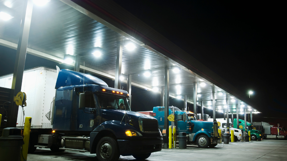 Varios camiones articulados estacionados bajo la marquesina iluminada de una estación-service de nuit, con un cielo oscuro de fondo, transporte internacional de mercancías.