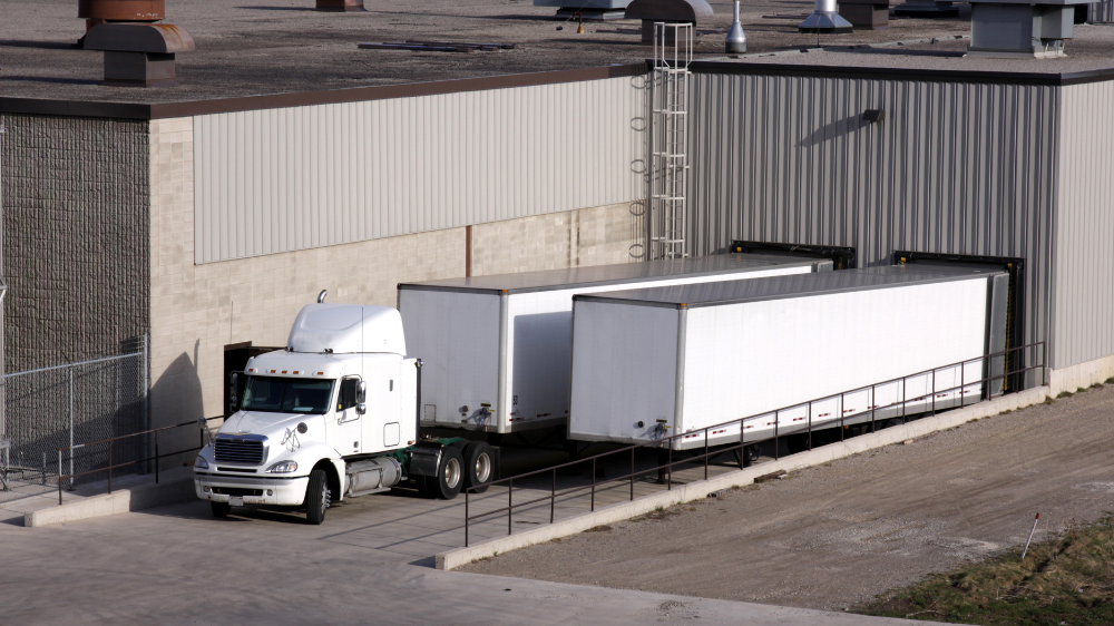 Camión blanco con doble remolque en los muelles de un almacén, representando flujos logísticos digitalizados mediante programas de gestión de transporte.