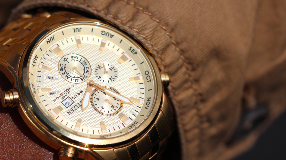 Primer plano de un reloj de pulsera metálico dorado con esfera blanca texturizada, tres subesferas, ventana de fecha y marcadores de mes, llevado sobre una manga marrón.