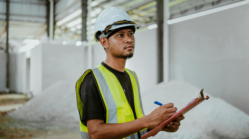 Ouvrier en casque blanc et gilet jaune inspectant des piles de matériaux dans un entrepôt industriel dans le cadre du suivi bennes chantier.
