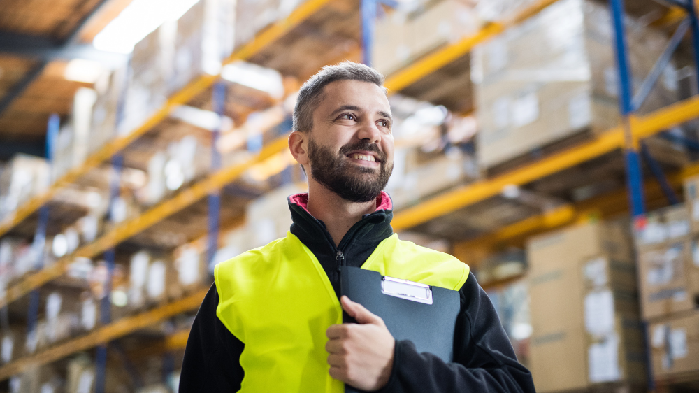 Employé en gilet haute visibilité souriant, tenant un clipboard dans un entrepôt aux rayonnages remplis de cartons, illustrant la gestion et la vérification d’une preuve de livraison en environnement logistique.