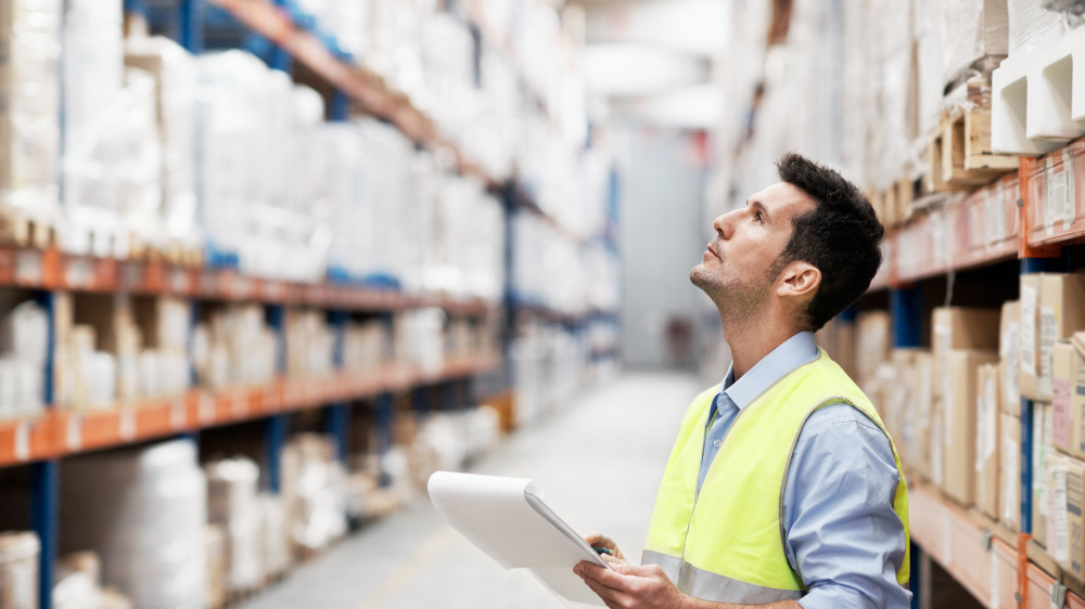 Homme en gilet de sécurité tenant un clipboard, observant un grand entrepôt rempli d’étagères avec des cartons et produits, afin de contrôler le stock minimum.