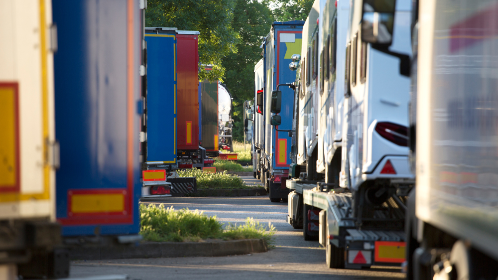 Vista entre remolques estacionados que muestra filas de camiones de carga de colores en un patio iluminado por el sol, con árboles al fondo, utilizada para contextualizar la gestión de horas de conducción.