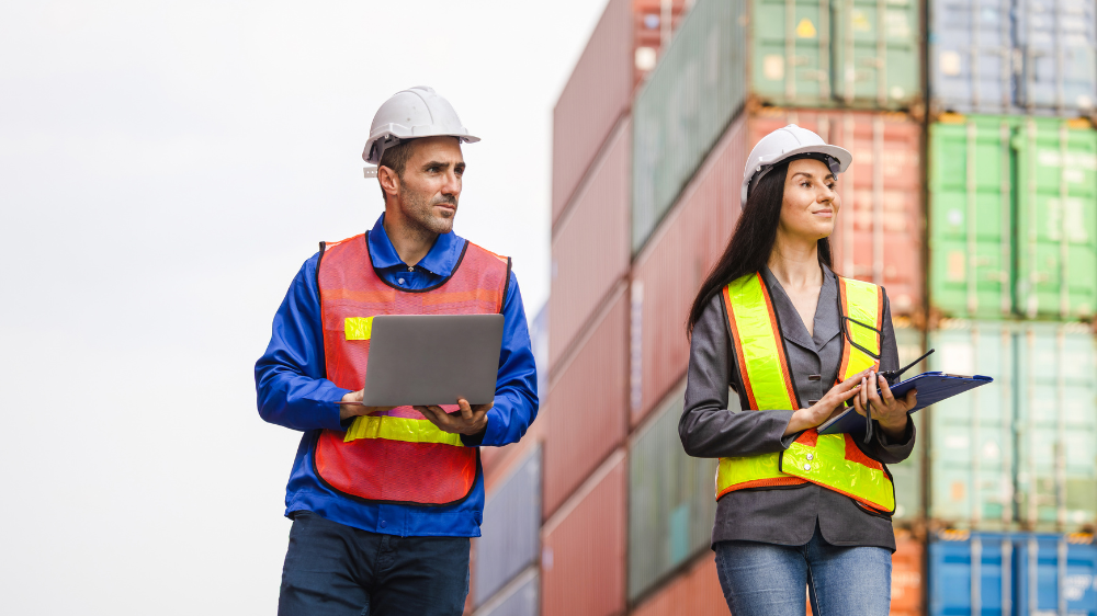 Trabajador de almacén con polo azul y gorra, de brazos cruzados frente a estanterías, representando procesos optimizados con programas de gestión de transporte.