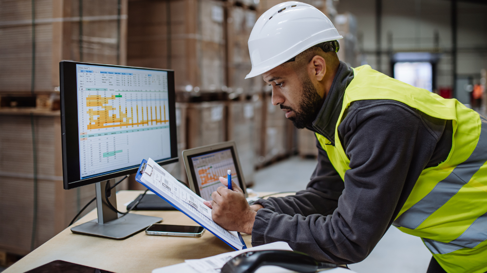 Homme en casque et gilet de sécurité écrivant sur une tablette dans un entrepôt de groupage, avec des écrans d’ordinateur affichant des données logistiques en arrière-plan.