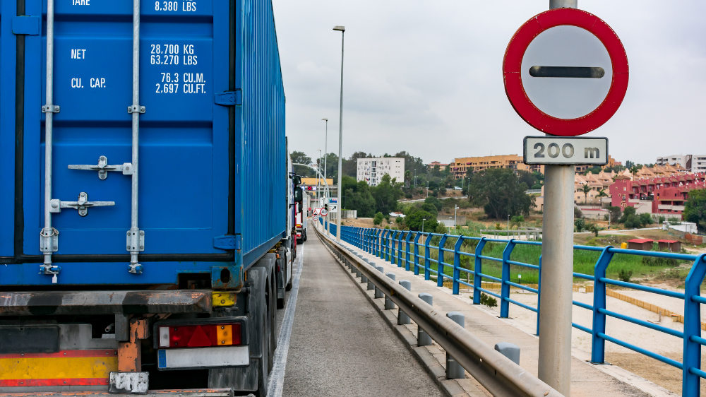 Camión azul sobre un puente estrecho con una señal de prohibido el paso para vehículos de más de 2,8 metros, con edificios visibles al fondo, transporte internacional de mercancías.
