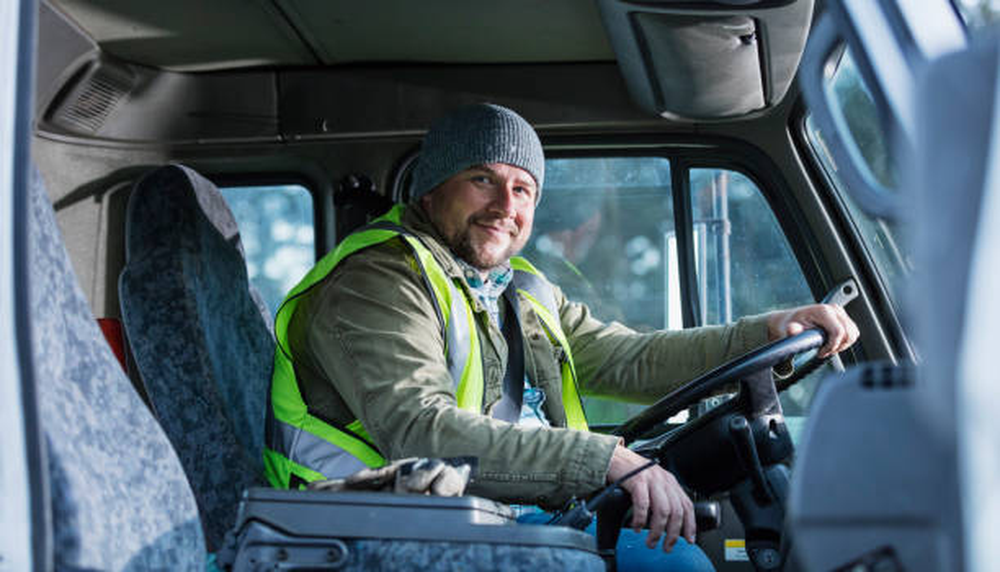 Hombre con gorro y chaleco de seguridad sonriendo mientras está sentado en el asiento del conductor de un camión con GPS telemático.