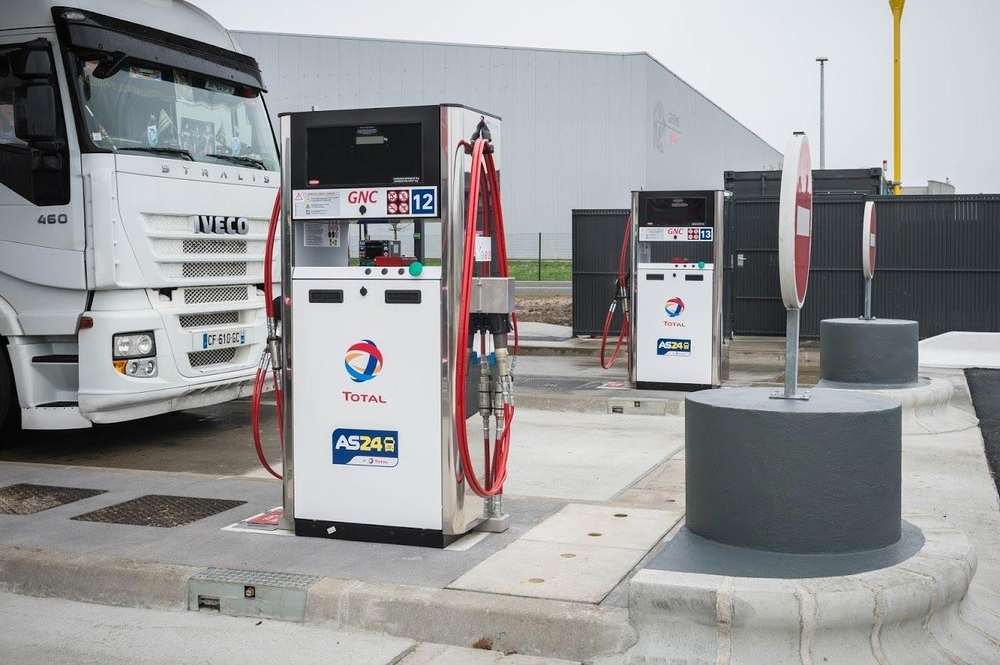 Station-service avec deux pompes à carburant, tuyaux rouges et un camion à proximité pour le remboursement TICPE. La station affiche les enseignes Total et AS24 dans une zone industrielle.