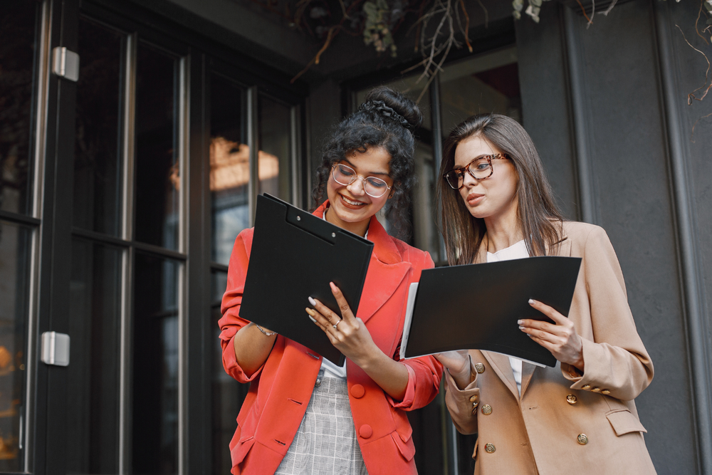 carta de porte ADR – dos mujeres revisando documentos en portapapeles al aire libre, con chaquetas rojas y beige, sonriendo