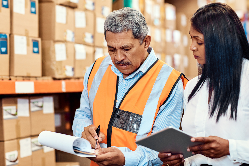 Dos operarios de almacén revisan documentos y una tableta en una zona de almacenamiento llena de cajas apiladas, ilustrando procesos de supply chain.