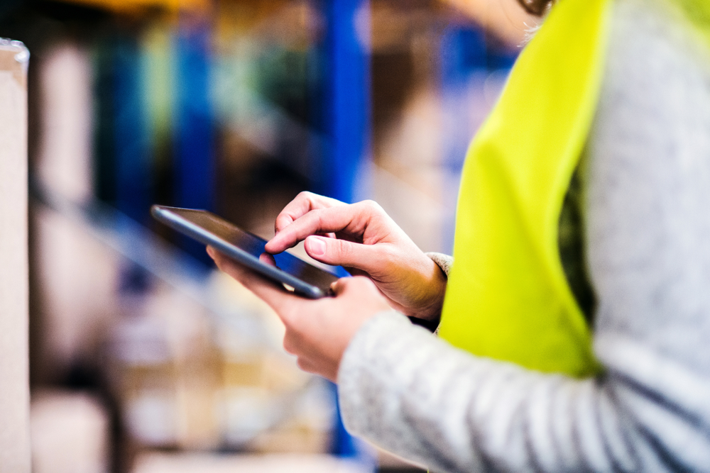 Personne en gilet jaune utilisant une tablette dans un entrepôt, entourée d’étagères et de cartons — une scène typique des opérations logistiques sous Incoterm CPT.