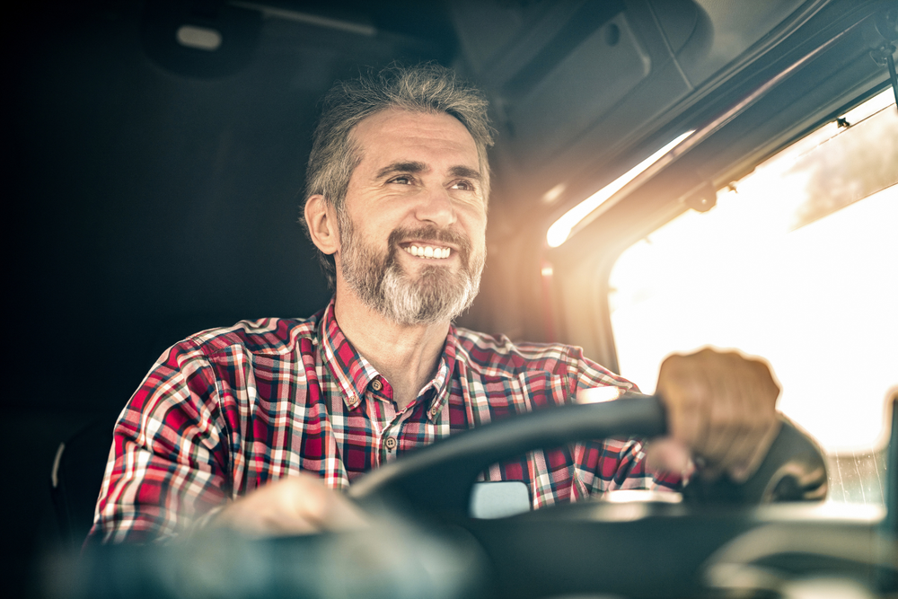 Hombre con barba y camisa de cuadros rojos sonriendo mientras conduce un vehículo, con el sol entrando por la ventana y el tacógrafo visible.