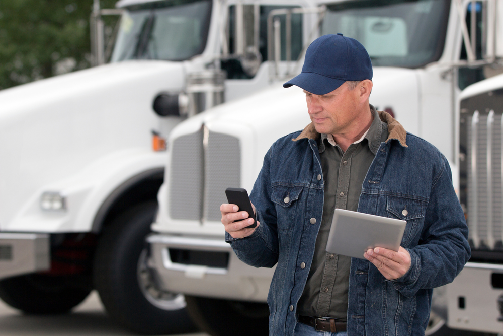 Homme en veste en jean et casquette devant des camions, tenant une tablette et consultant son téléphone, illustrant la carta de porte digitale.