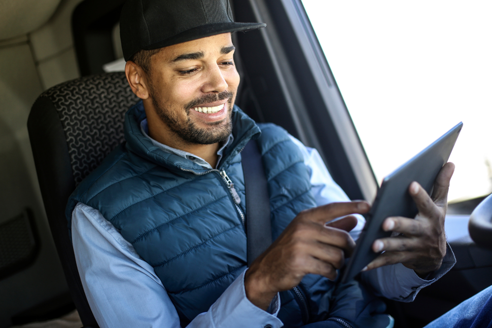 Hombre sonriente con gorra y chaleco azul usando una tablet desde el asiento del conductor de un vehículo, consultando un documento de transporte.