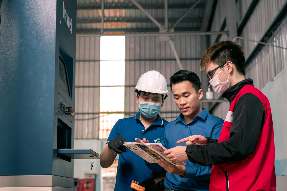 Trois travailleurs dans une usine discutant d’un document : l’un porte un casque, un autre un masque et le troisième tient le papier, coordination des créneaux horaires de production.