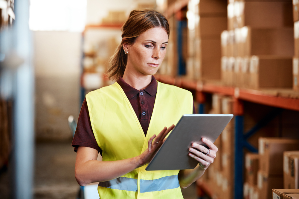 Mujer con chaleco amarillo de seguridad usando una tablet en un almacén con estanterías llenas de cajas, ilustrando la supply chain en la gestión logística.