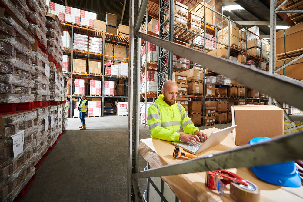 Employé en veste haute visibilité utilise un ordinateur portable dans un entrepôt 3PL logistique, entouré d’étagères de cartons et d’équipements, avec un autre travailleur en arrière-plan.