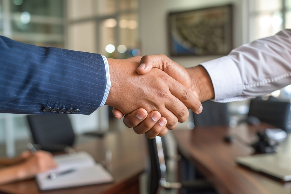 Dos personas se dan la mano en un entorno de oficina, con una mesa de reuniones y sillas desenfocadas al fondo — una colaboración facilitada por un sistema ERP que conecta equipos y procesos.