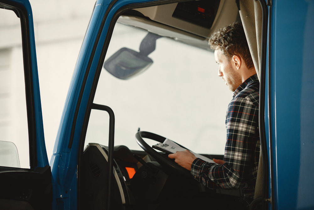 HMan in plaid shirt sitting in a blue truck's driver's seat, reading a document, with sunlight streaming through the window.