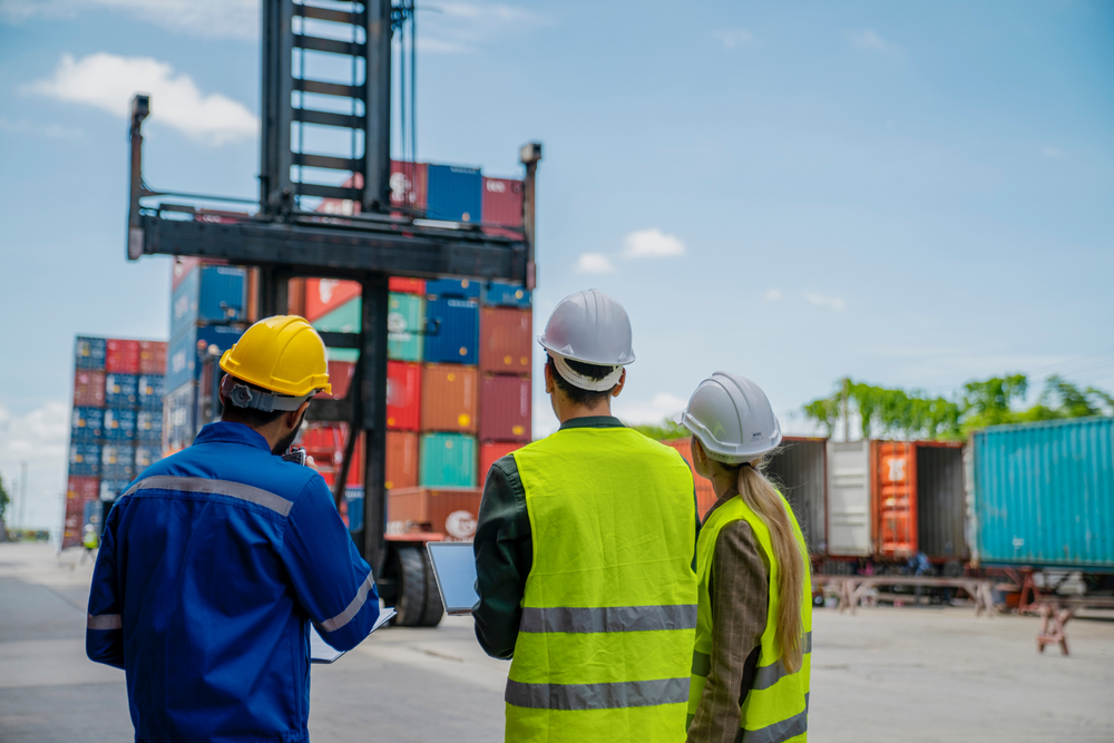 Tres trabajadores con equipo de seguridad observan una carretilla elevadora moviendo contenedores en un puerto, ilustrando la supply chain en operaciones de transporte y logística internacional.
