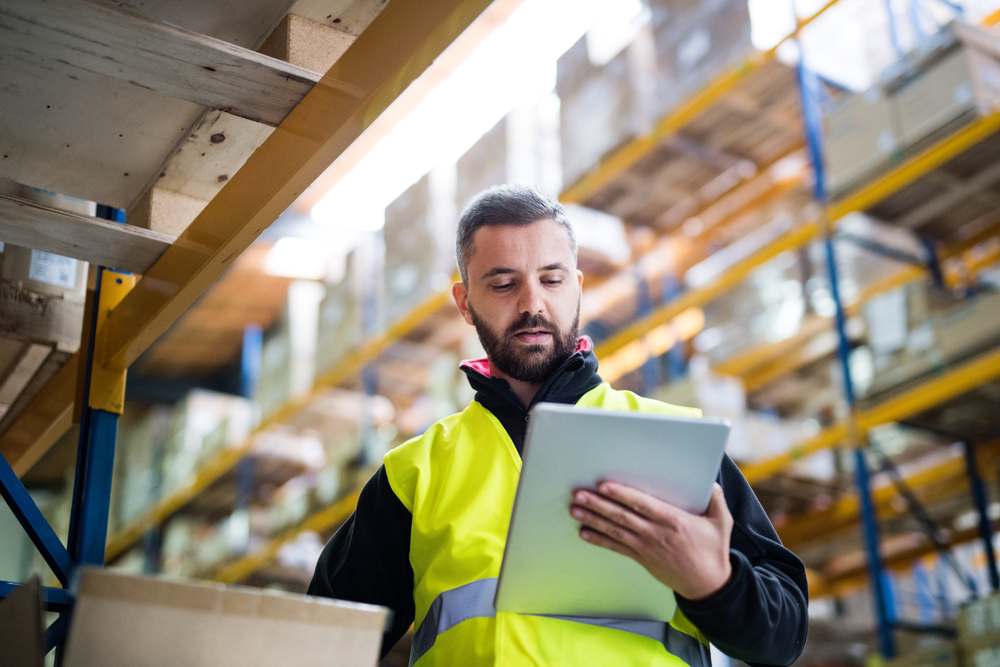 carta de porte ADR – trabajador de almacén con chaleco amarillo usando una tablet para gestión de inventario, rodeado de estanterías con cajas