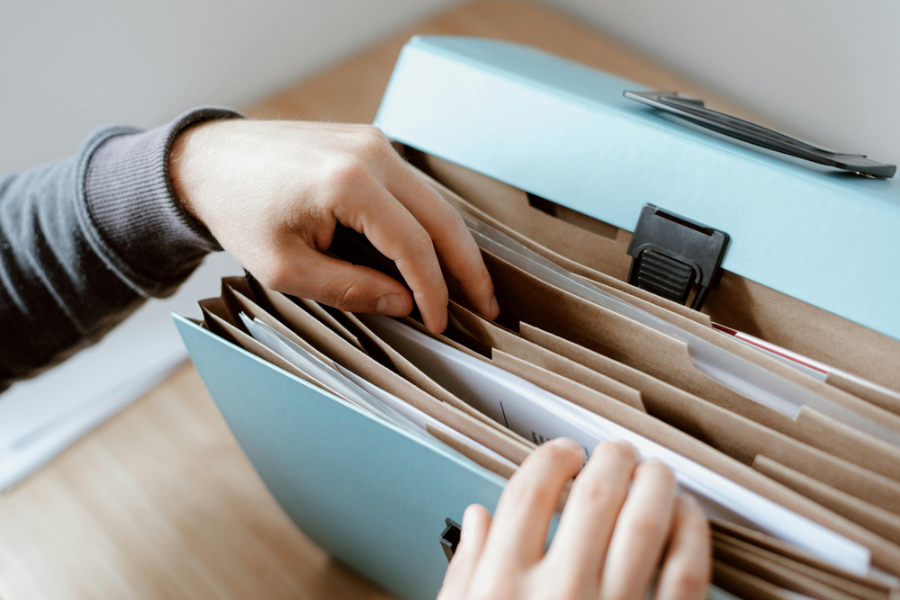 Mains organisant des documents de transport dans un trieur bleu à soufflet, posé sur une surface en bois.        Demander à ChatGPT