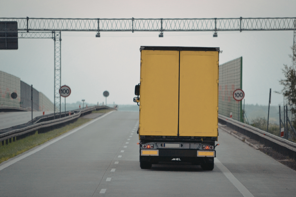 Camión de reparto amarillo circulando por autopista vacía con señal de límite 100 km/h y cielo nublado al fondo.
