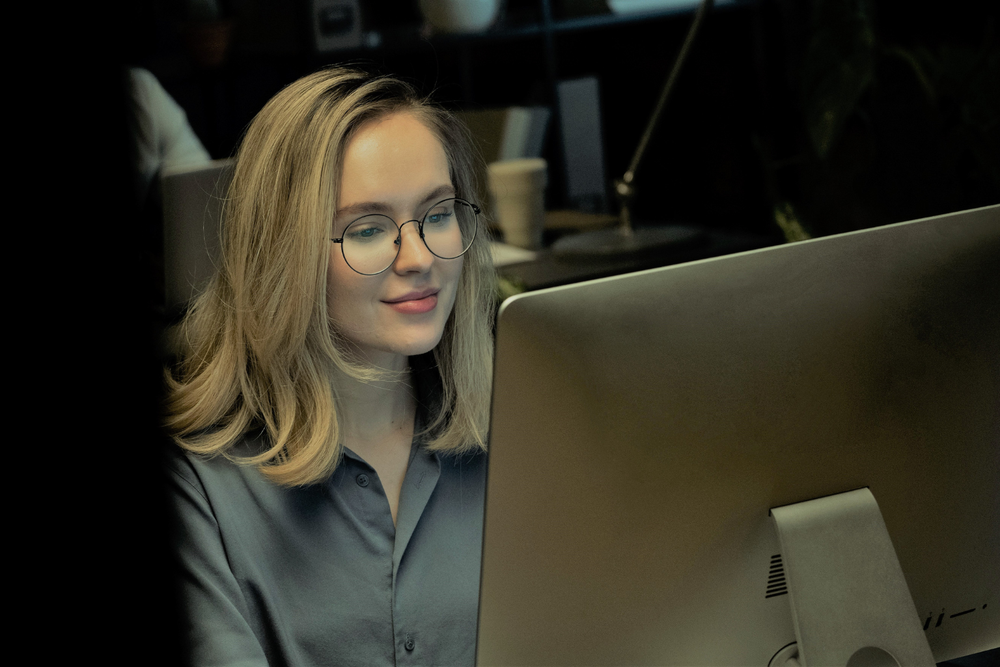 Femme blonde portant des lunettes travaillant sur un ordinateur dans une pièce faiblement éclairée, illustrant la gestion numérique des données pour décarboniser le transport.
