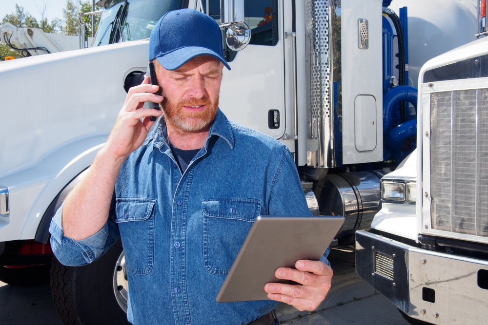 Homme en chemise en jean et casquette utilisant une tablette et parlant au téléphone, debout entre deux camions, dans le cadre d’une opération logistique conforme à l’Incoterm CPT.