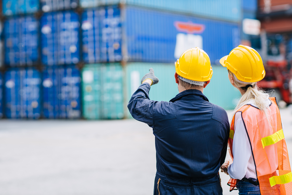 Deux travailleurs en tenue de sécurité et casque pointent des conteneurs empilés dans une zone de fret, sur un site logistique illustrant l’application de l’Incoterm CPT.