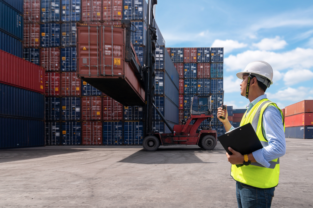 Un opérateur en casque et gilet de sécurité supervise avec un clipboard et une radio le déplacement de conteneurs par un chariot élévateur dans une zone de stockage, illustrant la coordination logistique entre WMS TMS.