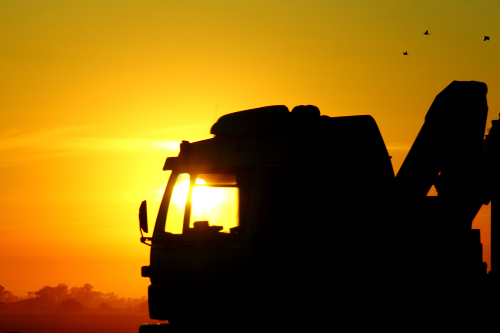 Silueta de camión de transporte al amanecer con cielo naranja y amarillo y aves volando.