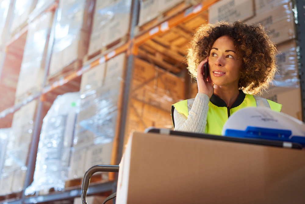 Femme en gilet haute visibilité parlant au téléphone dans un entrepôt, entourée de cartons empilés et tenant un bon de livraison