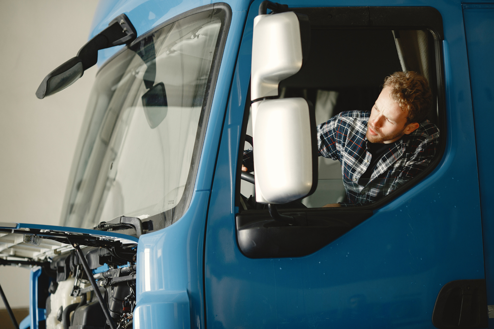 Hombre con camisa de cuadros se asoma por la ventana de un camión azul, revisando el retrovisor y el motor abierto, antes de completar la carta de porte.
