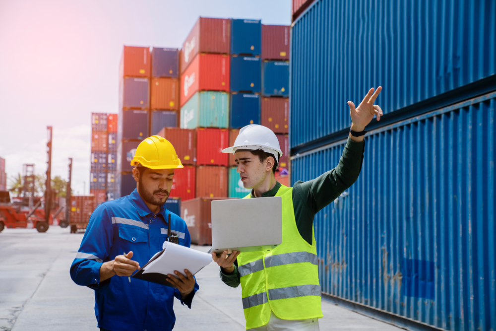 Deux travailleurs en tenue de sécurité discutant logistique dans une cour de fret entourée de conteneurs d’import export