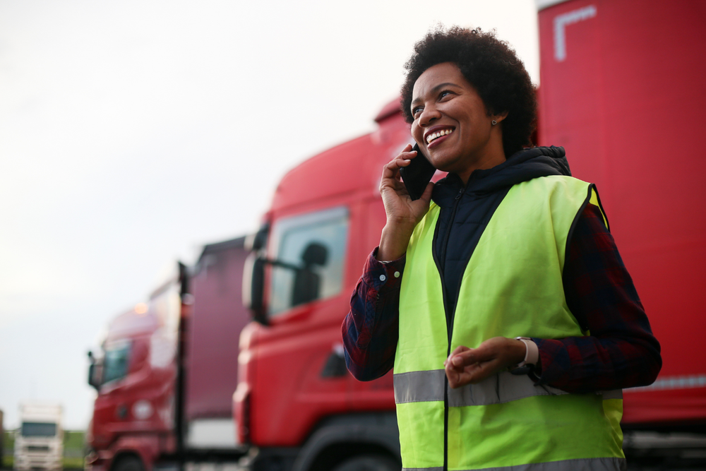 Une personne souriante en gilet réfléchissant parle au téléphone devant des camions rouges, symbole de coordination et de performance dans l’externalisation logistique.