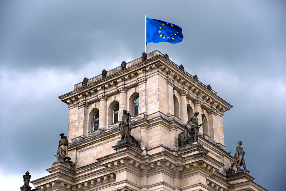 Bandera de la Unión Europea ondeando sobre un edificio de piedra con estatuas bajo un cielo nublado