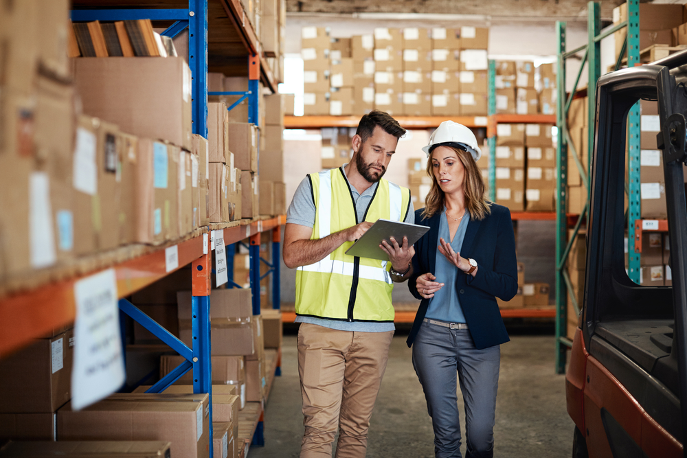 Deux personnes en entrepôt discutant du flux tendu en logistique avec un clipboard parmi des étagères de cartons.