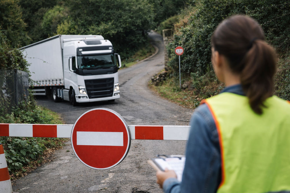 Trabajador con chaleco reflectante sosteniendo un portapapeles bloquea una carretera con una señal de prohibido el paso, deteniendo un camión en una vía rural por una orden de transporte no conforme