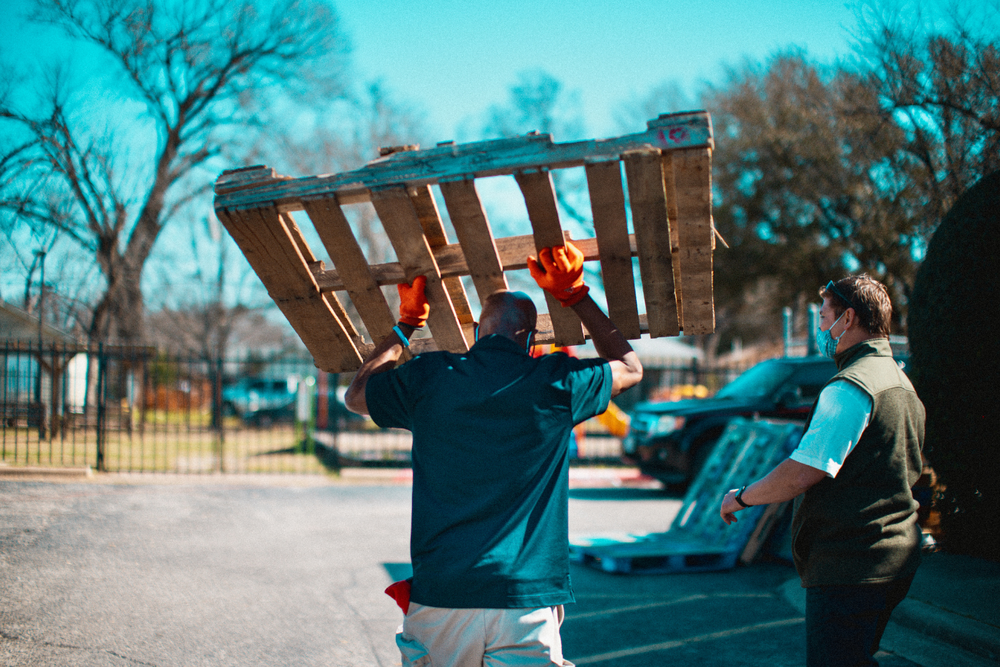 Dos hombres al aire libre, uno cargando un pallet de madera, ambos con guantes, con árboles y una valla al fondo.