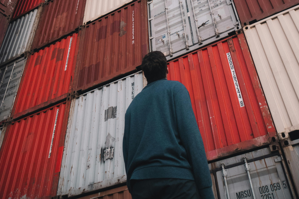 Personne en pull bleu observant des conteneurs rouges et blancs empilés dans une zone d’import export