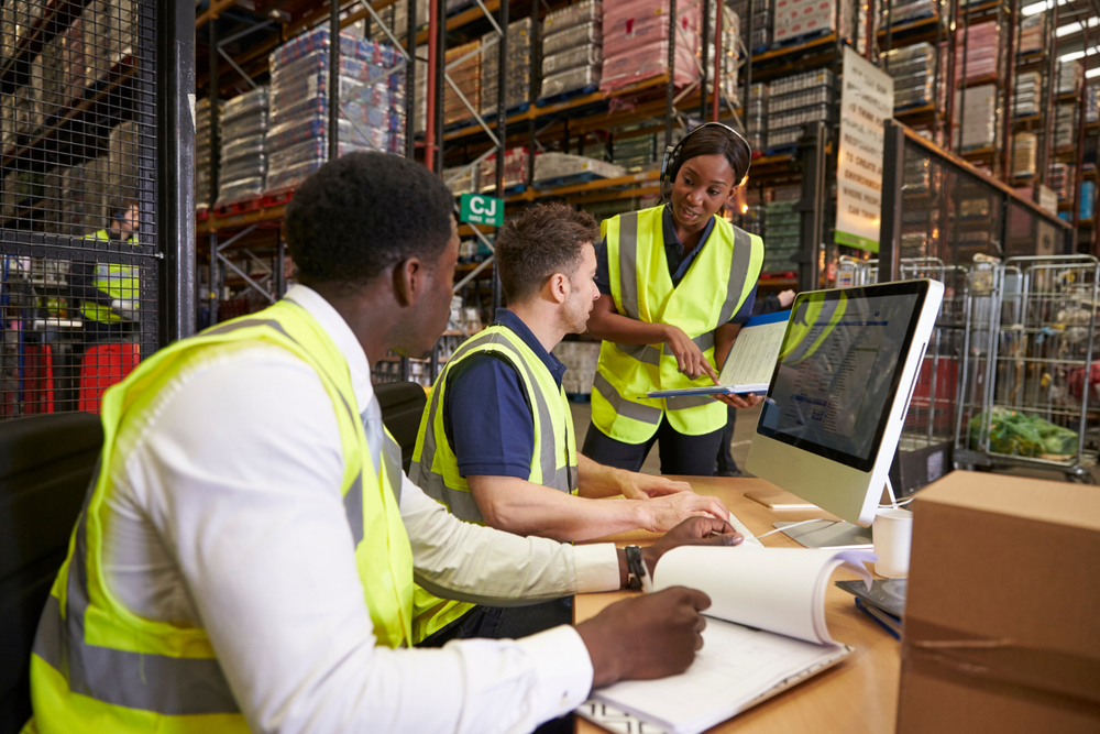 Équipe logistique en gilets jaunes analysant les processus de flux tendu en logistique sur un ordinateur devant des rayonnages de stockage remplis de cartons.