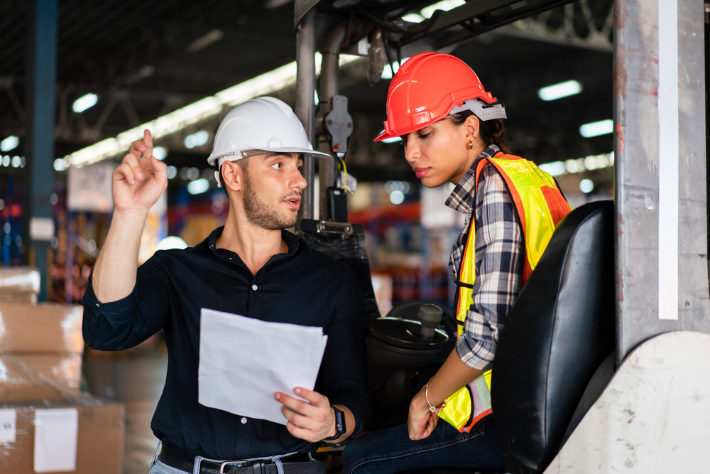 Homme et femme en casque de sécurité discutant d’un document dans un entrepôt avec TMS chargeur.