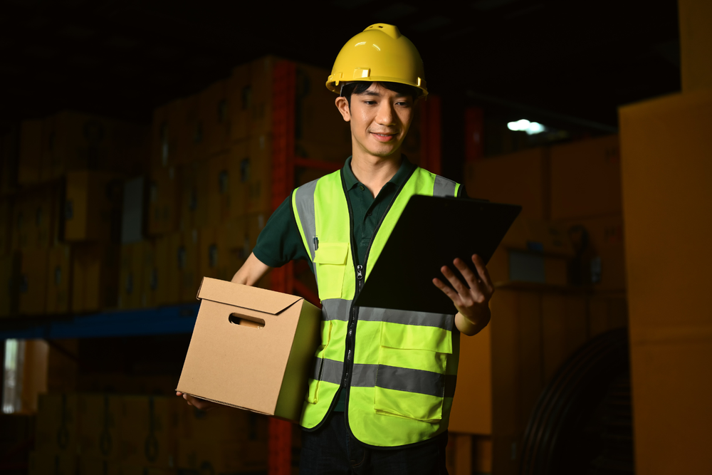 Un operario de almacén con casco amarillo y chaleco reflectante sostiene una caja mientras revisa una hoja en una carpeta, rodeado de cajas de cartón apiladas — todo gestionado mediante un sistema ERP que centraliza la operativa logística.