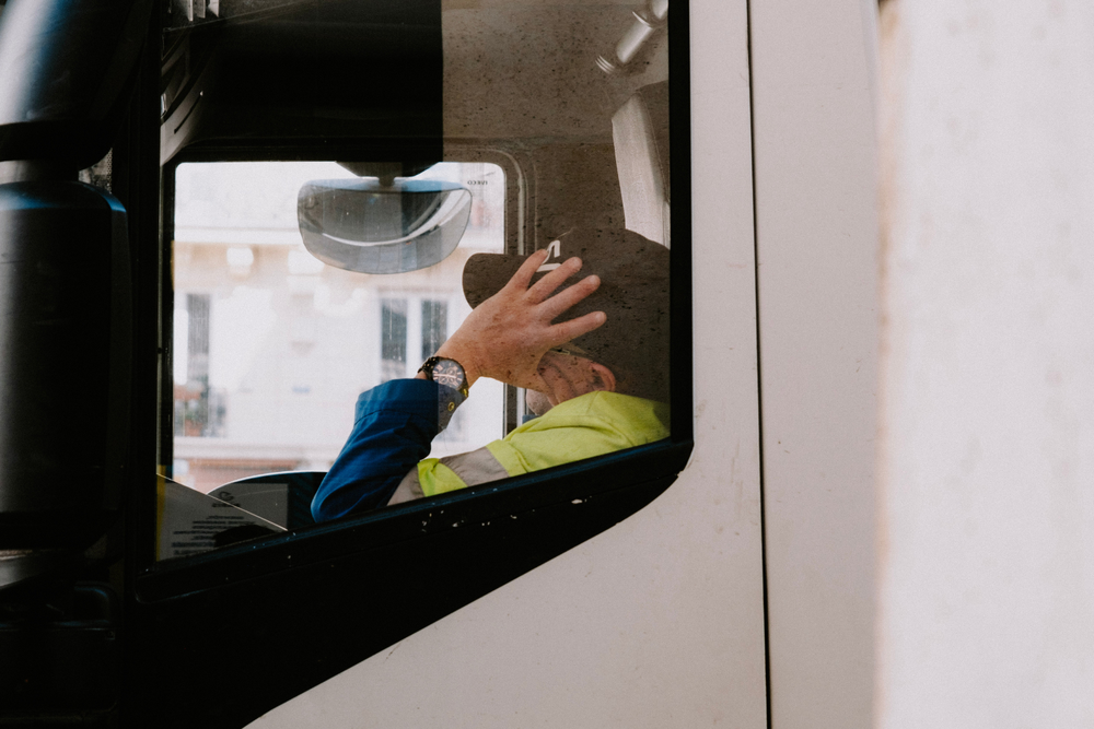 Conductor de camión con chaleco de seguridad amarillo, visto a través de la ventana, sostiene un cigarrillo y se toca la cabeza mientras está sentado en la cabina