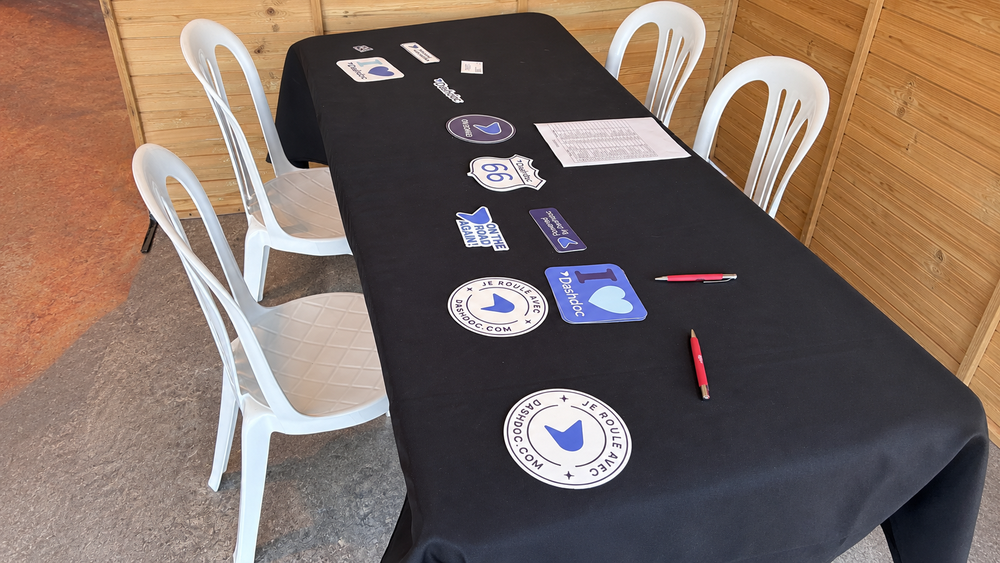 Table à nappe noire et chaises blanches avec stickers (motifs bleus, panneaux routiers) et stylos, sur le stand de l'AG France Plateaux 2026