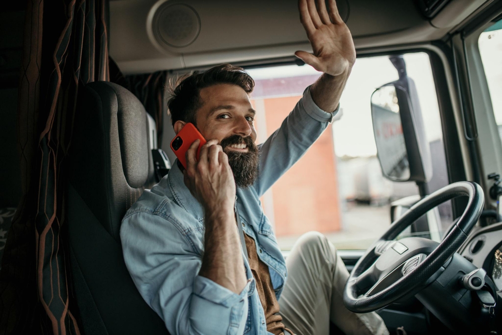 Hombre dentro de la cabina de un camión, sonriente, hablando por teléfono rojo y saludando con la mano por la ventana abierta escena cotidiana del transporte vinculada al uso del gasóleo profesional.
