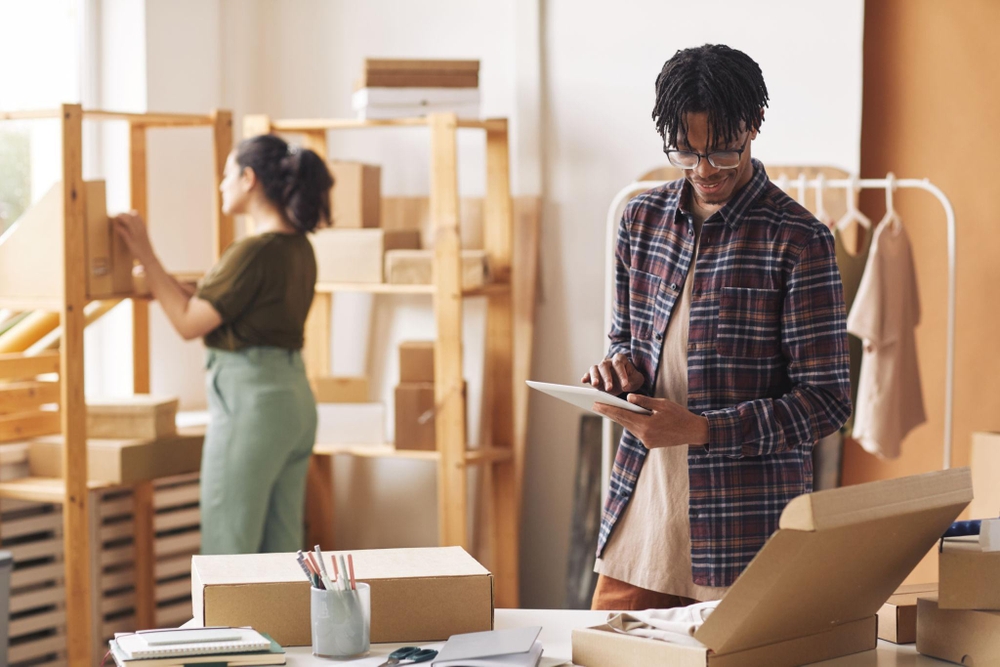 Deux personnes gèrent la logistique commerce dans un espace de travail, organisent des commandes avec une tablette, entourées de cartons et d’étagères.