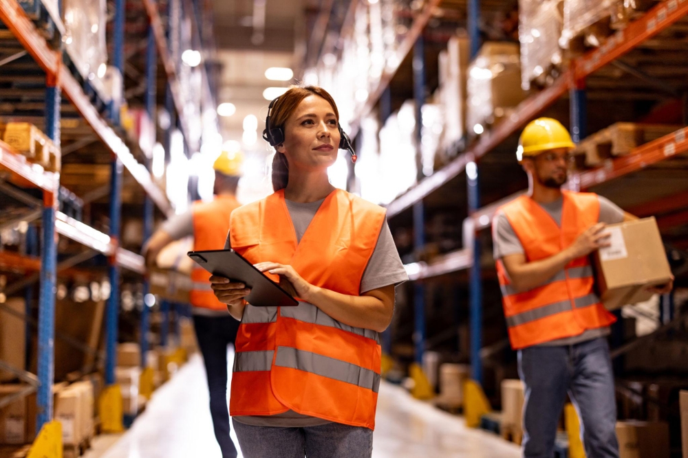 Opérateurs en entrepôt en gilets orange et casques gèrent la logistique commerce : une femme utilise une tablette en marchant dans une allée bordée de rayonnages remplis de cartons.
