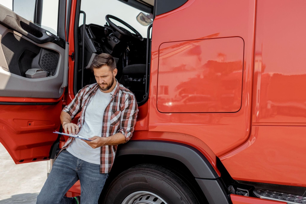 Hombre apoyado en un camión rojo con la puerta del conductor abierta, sosteniendo un portapapeles y vestido con camisa de cuadros y vaqueros — escena operativa del día a día en transporte vinculada al seguimiento del gasóleo profesional.